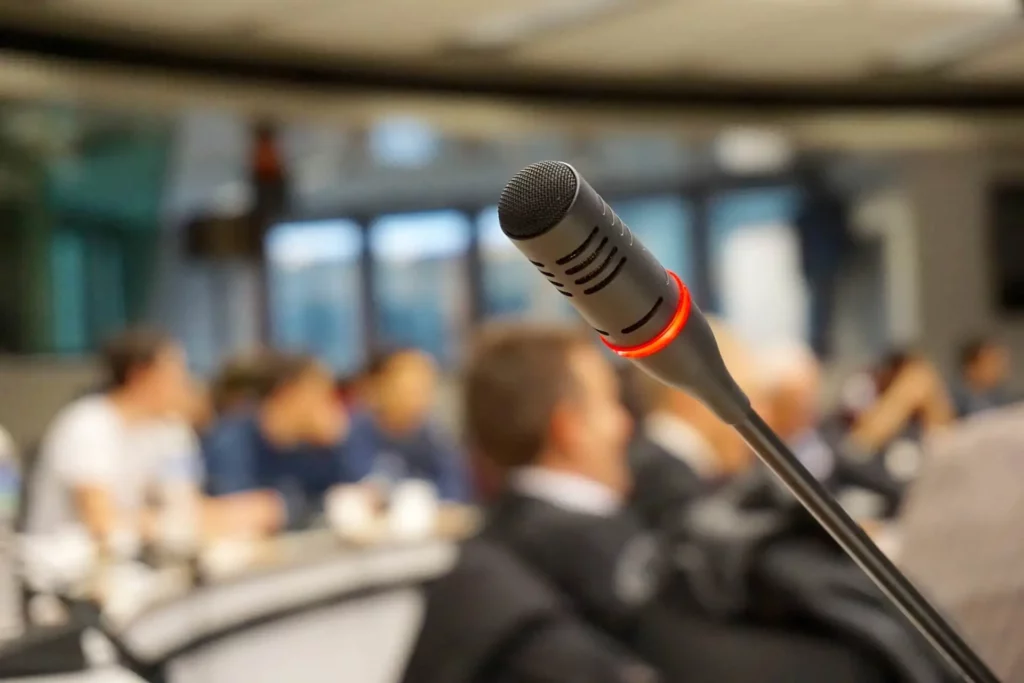 Ein Konferenzmikrophon im Vordergrund, im Hintergrund unscharf Teilnehmer einer Konferenz.
